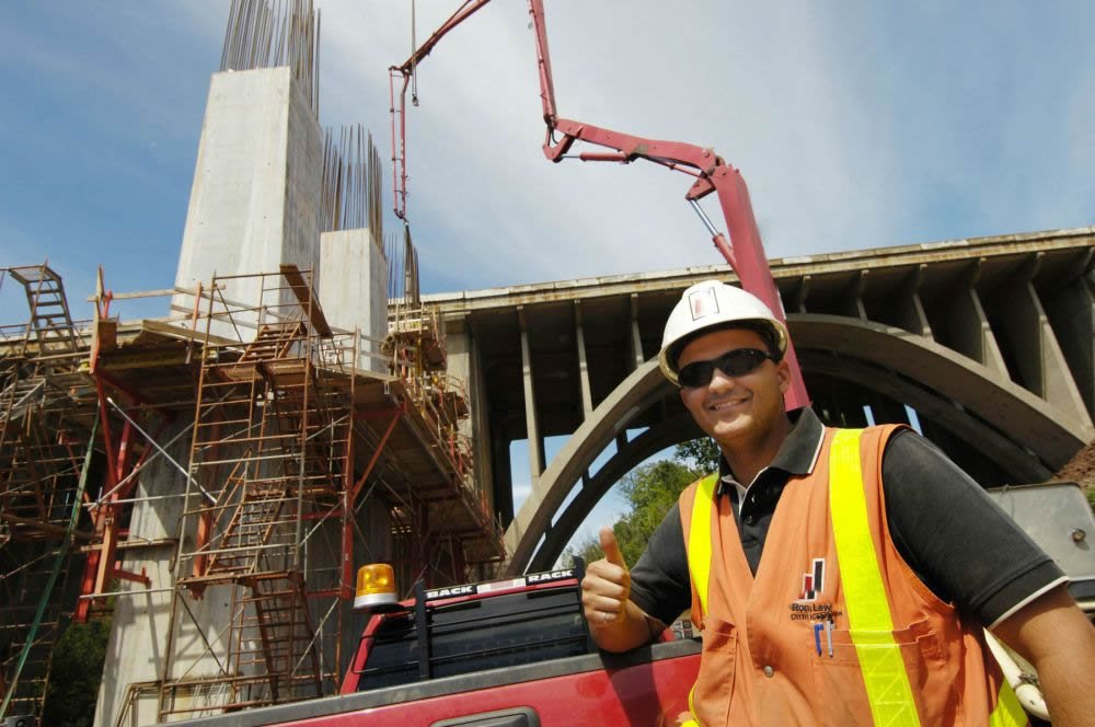 Superintendent with thumb up standing in front of a pickup truck with a concrete pump truck in the distance pouring a column.